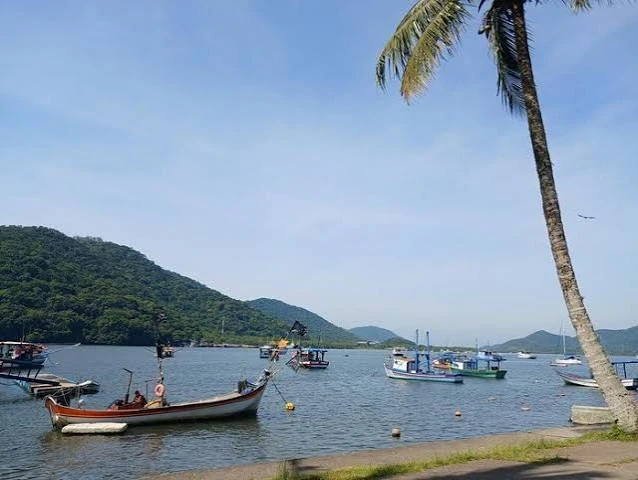 A peaceful, moody landscape of a wide, dark brown river flowing smoothly between densely forested, green banks. The water reflects an overcast, dusky sky. A small, silver rowboat is moored on the grassy bank in the lower right foreground.