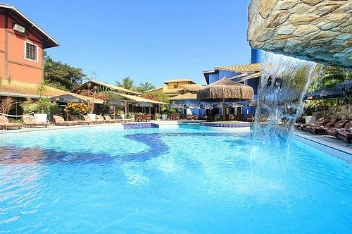 An expansive outdoor swimming pool with a large artificial rock formation that features a cascading waterfall, surrounded by white lounge chairs and colorful resort buildings with thatched roofs under a clear blue sky.