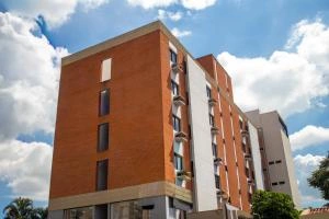 Angled exterior view of a contemporary mid-rise apartment or hotel building, featuring vertical sections of red brick alongside white stucco walls and narrow glass windows, set against a bright blue sky with fluffy white clouds.