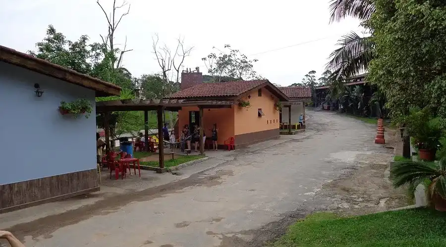 View of a concrete road passing through a rural or countryside area, lined with a light blue house and a peach-colored house featuring a red clay tile roof and a covered patio with an outdoor seating area and red plastic chairs.