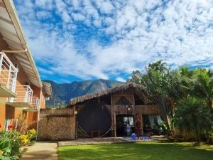 A beautiful daytime view of a resort courtyard featuring a large, thatched-roof open-air pavilion, surrounded by green grass and tropical palm trees. In the background, towering green mountains rise against a bright blue sky filled with textured white clouds.