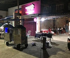Nighttime outdoor street food setup featuring metal smokers, a bright pink glowing storefront sign, and patrons seated at outdoor tables.