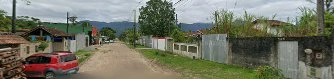Unpaved dirt road running through a residential neighborhood, lined with fenced houses, green trees, and mountains in the background.