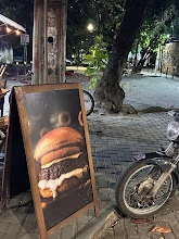 Wooden A-frame chalkboard displaying a large burger image, placed on a brick sidewalk next to a tree and a parked motorcycle at night.