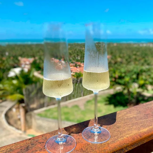 Two condensation-covered champagne glasses filled with bubbly white wine, resting on a rustic wooden railing with a tropical ocean and palm tree landscape in the background.
