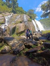Three people standing and sitting on large, flat boulders at the base of a rocky, multi-stream waterfall. Other hikers can be seen exploring the surrounding rocks, with bright sunlight and clear blue skies above.