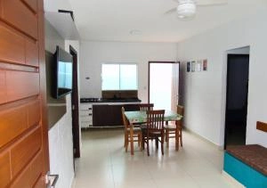 Interior view of a clean, modern, open-concept room with white walls and a white ceiling fan. The space includes a wall-mounted flat-screen television, a wooden dining table with four chairs, a dark kitchen counter in the background, and an open doorway leading outside.
