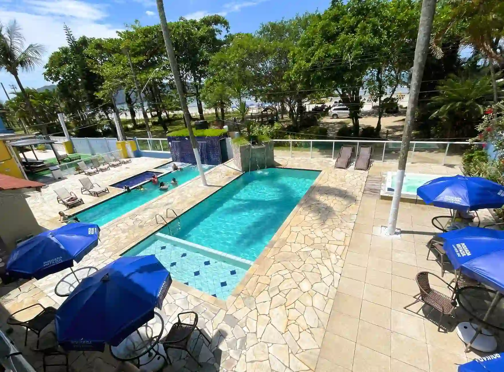 Sunny outdoor swimming pool area at a resort featuring rectangular blue pools, a stone patio with lounge chairs, several bright blue umbrellas, and a view of dense green trees and the sandy beach in the background.
