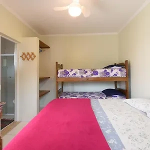Interior view of a bright bedroom featuring a wooden bunk bed set and a single bed, both dressed with purple and white floral bedding, alongside a ceiling fan and corner shelving.