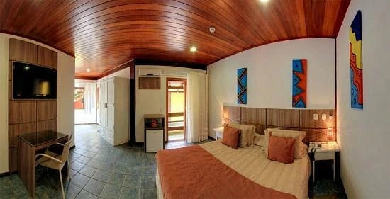 Interior view of a spacious hotel room featuring a stunning arched ceiling made of polished wooden planks, a large bed with brown and orange pillows, a flat-screen TV on the wall, and colorful abstract art.
