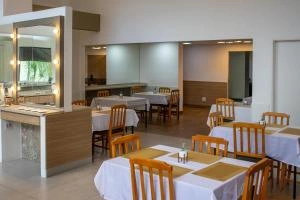 A spacious and bright dining area for guests, featuring multiple tables neatly arranged with white tablecloths, tan placemats, and wooden chairs, alongside a built-in light wood buffet station with a mirrored backsplash.