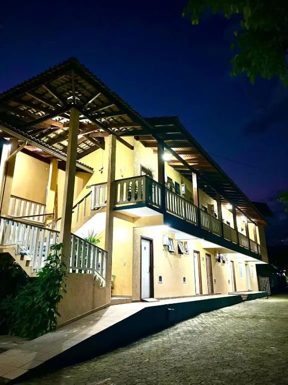 An exterior night shot of a two-story yellow guesthouse or hotel with a dark wooden balcony and a traditional sloped roof. Bright exterior lights illuminate the walkway, stairs, and individual room doors against a deep blue night sky.