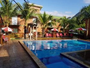 A vibrant resort pool scene showing a rectangular blue swimming pool situated on a wooden deck. The area is surrounded by lush palm trees, a wooden fence, and several red patio umbrellas providing shade over lounge chairs, with multi-story red resort buildings in the background.