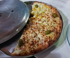 Sliced cheese and ham pizza topped with green olives and corn, served on a metal tray with a partial metal lid cover.