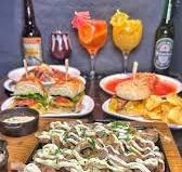 Assorted food platter including sliders with fries, grilled meat chunks with creamy sauce, mixed cocktails, and beer bottles on a dark background.
