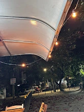 Nighttime view of outdoor patio seating under a clear plastic awning, illuminated by warm string lights, with street trees and cars in the background.