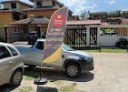 Tall standing promotional feather flag with a red and yellow logo, placed on a gravel lot in front of parked cars and a gated residential property.