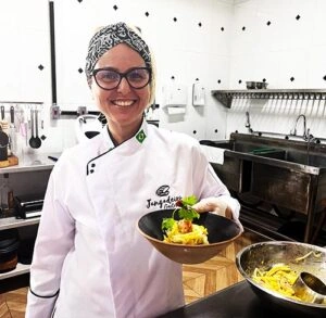 Smiling female chef wearing a white chef coat and patterned headscarf, holding a black bowl containing a shrimp appetizer in a professional kitchen.