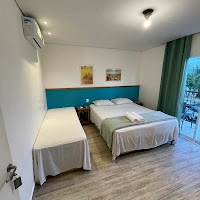 Exterior view of a multi-story beachfront Pousada hotel situated directly on the sand, featuring open ground-floor dining with umbrellas, balconies on upper floors, and lush green trees with mountains visible in the distance under a warm sunset or sunrise light.