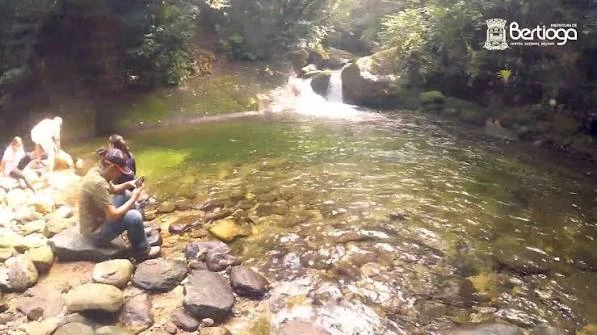 A group of people sitting on smooth stones along the edge of a clear, shallow river pool, with a small waterfall in the background and a "Bertioga" logo visible in the top right corner.