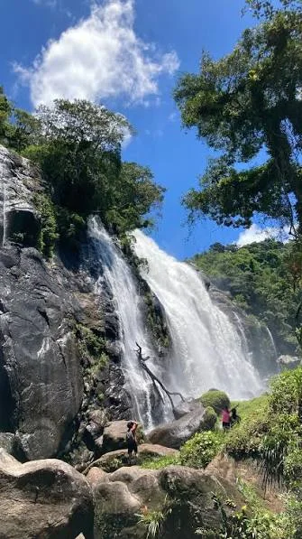 An upward-facing vertical shot of a majestic, wide waterfall flowing over dark rocky cliffs. A large dead branch is caught on the rocks, and tiny figures of hikers at the bottom provide a sense of the waterfall's massive scale against a bright blue sky with fluffy white clouds.