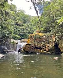 An elevated, aerial shot of a long, narrow waterfall flowing down the side of a mountain through dense, green forest, set against a backdrop of rolling hills, distant mountains, and a blue sky with scattered white clouds.
