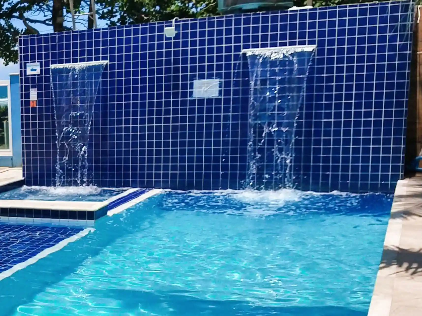 Close-up view of a resort swimming pool with vibrant blue water, featuring dual sheet waterfalls cascading from a striking wall of dark blue square mosaic tiles under natural daylight.