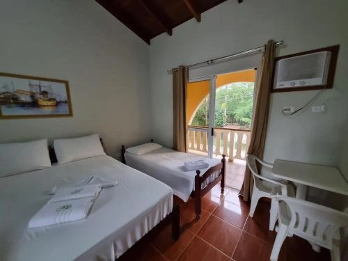 A bright guest bedroom featuring two beds with pristine white linens and neatly folded towels, a small white dining table with chairs, and large sliding glass doors with yellow arched framing that open to a balcony overlooking green trees. A framed painting of a boat hangs on the wall alongside a wall-mounted air conditioning unit.