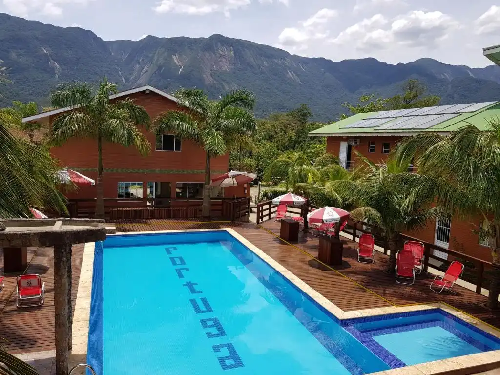 A sunny outdoor swimming pool featuring a blue-tiled bottom with the word "Portugal" spelled out in mosaic tiles. The pool is surrounded by a wooden deck, red lounge chairs, and red-and-white umbrellas, with resort buildings, tall palm trees, and a majestic, lush mountain range forming a stunning scenic backdrop.