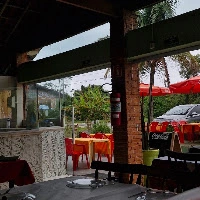 Open-air restaurant patio with glass enclosure, red plastic chairs, outdoor dining tables, red umbrellas, and parked cars visible.