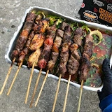 Metal tray filled with grilled chicken and beef skewers over fresh greens, held by a person wearing a black glove and a branded black apron.