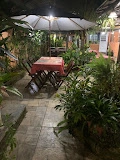Nighttime outdoor patio area with lush green plants, a white umbrella, and a wooden table covered with a red tablecloth.