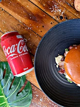 Top-down view of a burger on a black textured plate, placed next to a red Coca-Cola can on a wooden table scattered with salt and a green leaf.