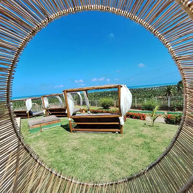 Two wooden daybeds with white drapes set on green lawn, framed perfectly through a circular woven bamboo structure, with blue ocean and sky in the distance.