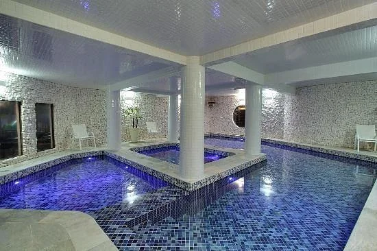 A relaxing indoor swimming pool area filled with blue mosaic tiles, surrounded by white square pillars and walls, with a circular jacuzzi section in the corner and white lounge chairs for resting.