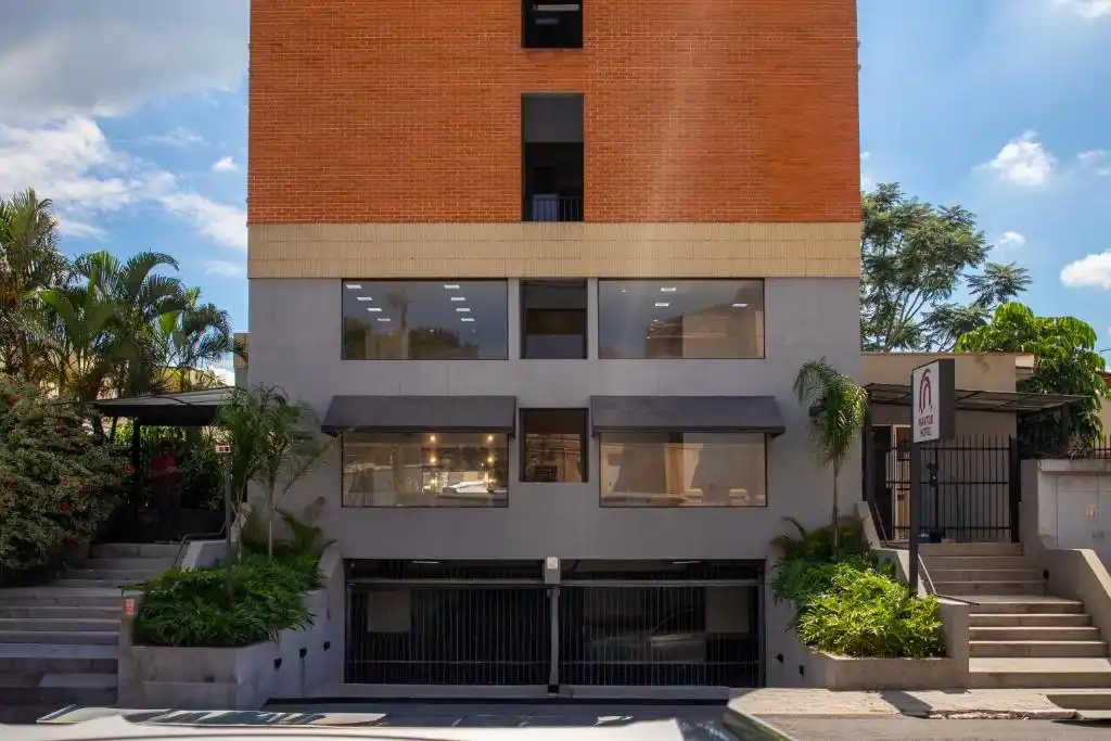 Facade of a modern hotel building featuring a red brick upper section and a gray lower level with large glass windows and black awnings. The street-level entrance includes a metal security gate, concrete steps, and lush tropical landscaping, with a vertical hotel sign visible on the right.