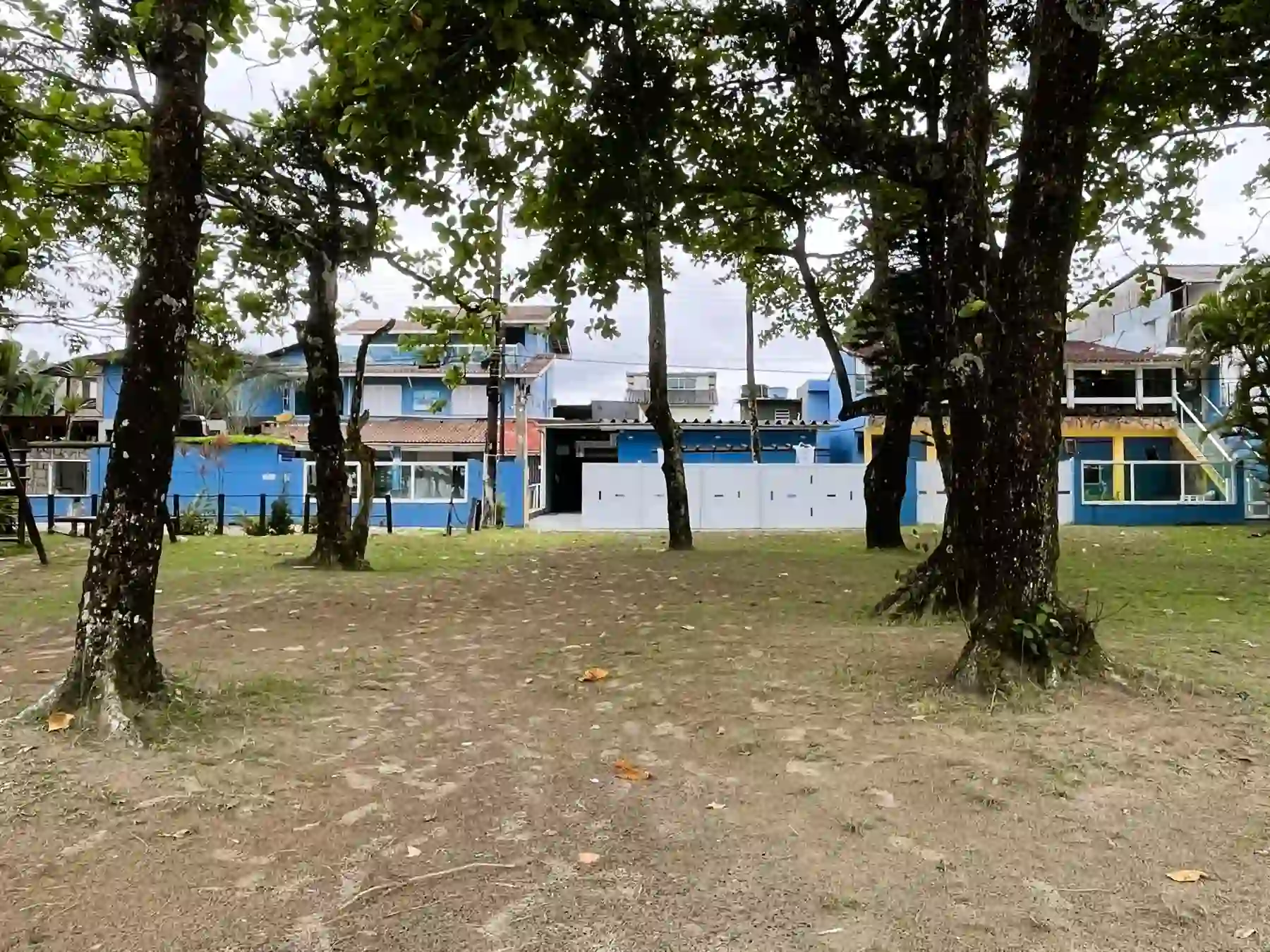 Tranquil outdoor courtyard scene showing a sandy and grass ground area framed by large, mature dark-barked trees, with a view of a white fence and vibrant blue multi-story buildings with balconies in the background.