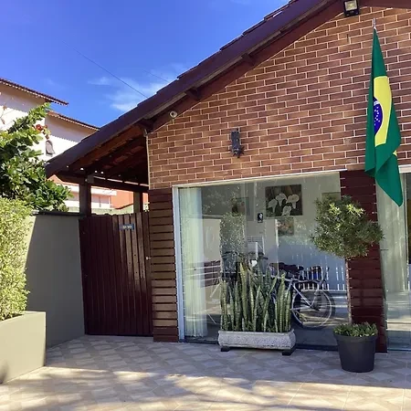 Exterior view of a modern red brick house with large sliding glass doors, a dark wood fence, and a Brazilian flag hanging on the wall, accented by potted cacti and an olive tree.