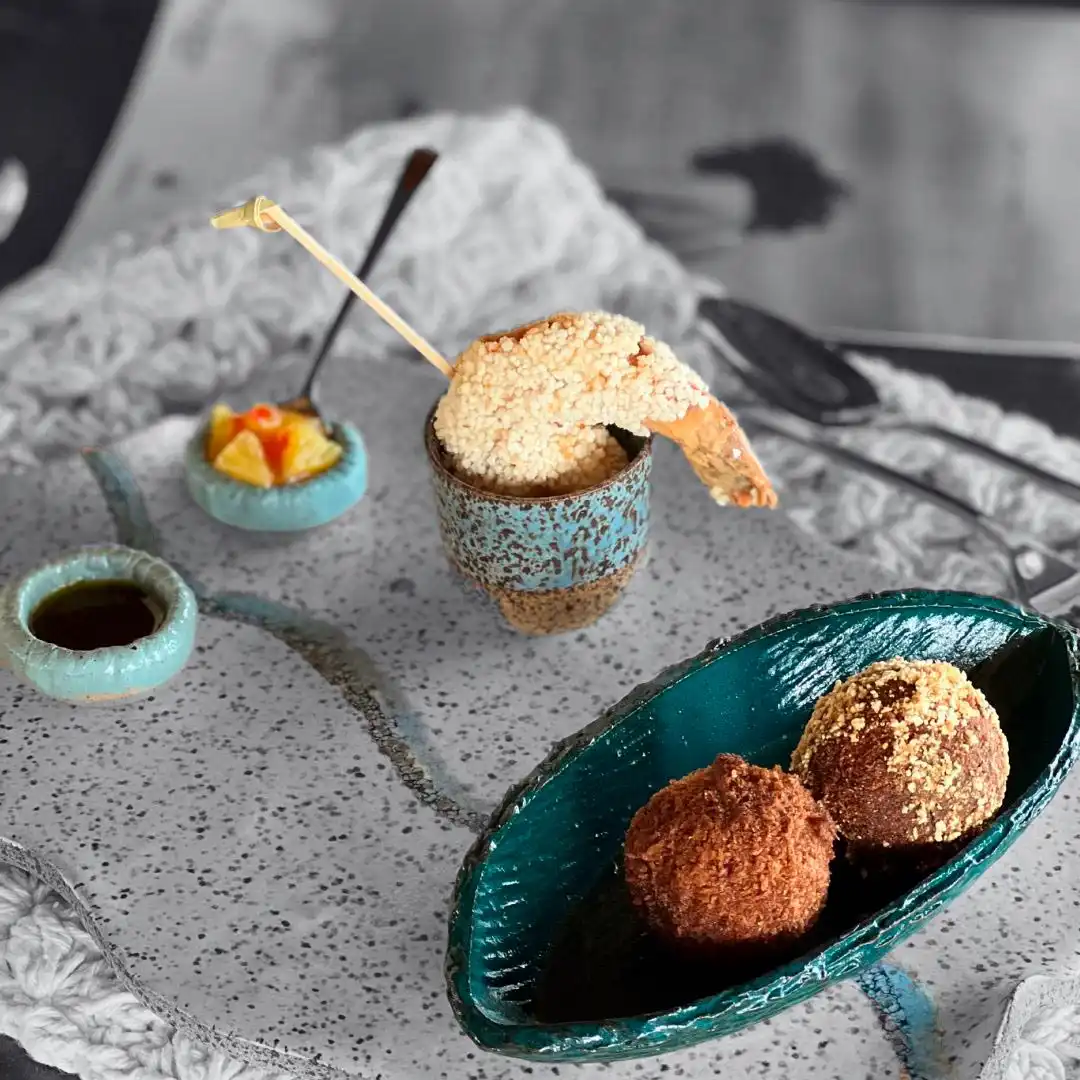 Gourmet appetizer arrangement featuring a sesame-crusted shrimp on a skewer, two fried croquette balls, and small bowls of diced fruit and dark sauce, styled on a textured surface.