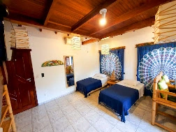 Intimate twin-bed guest room featuring deep blue bed runners, a rustic solid wood wardrobe, woven pendant lighting, and patterned blue-and-white drapes covering the window.