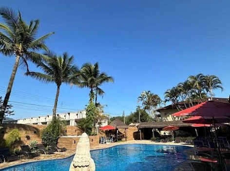 Sunny outdoor resort swimming pool with clear blue water, surrounded by a brick wall, tall palm trees, patio lounge chairs, and red umbrellas under a clear sky.