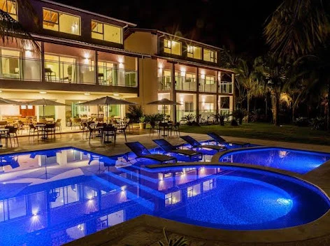 Illuminated outdoor swimming pool area at night featuring vibrant blue underwater LED lights, modern black sun loungers, and a multi-story hotel building with warm, lit balconies.