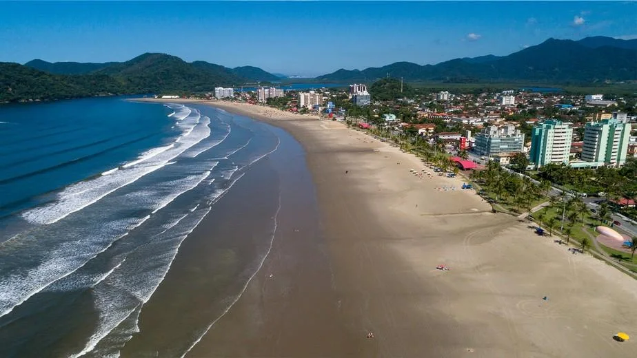 Aerial view of a sweeping crescent-shaped beach. The ocean features several parallel lines of white breaking waves, while a wide expanse of smooth, brown sand meets the water. Behind the beach, a coastal city with high-rise apartment buildings sits at the foot of lush, green, rolling mountains under a clear blue sky.