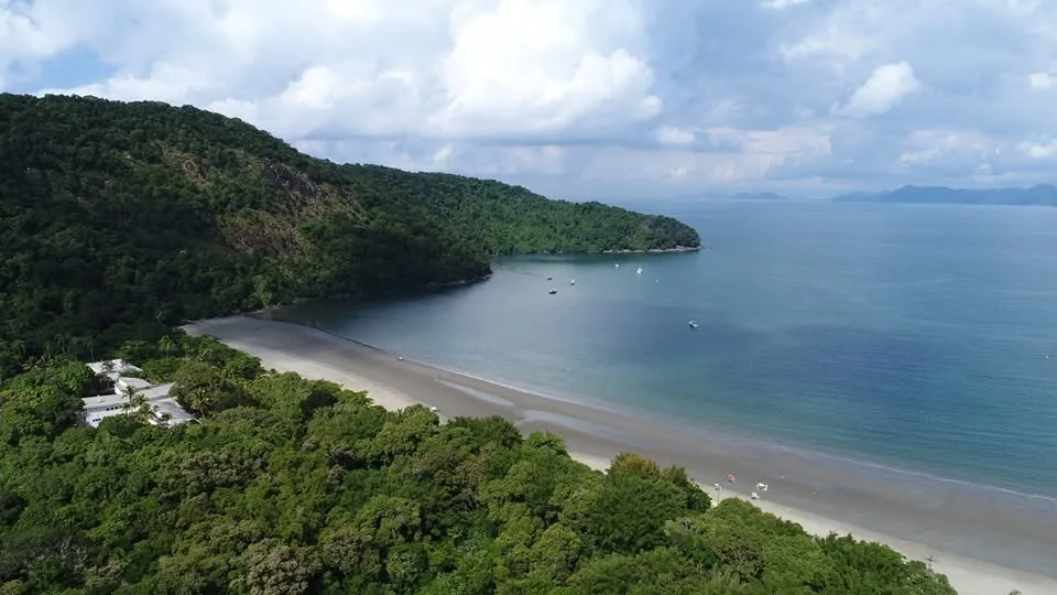 An aerial view of a secluded, crescent-shaped beach surrounded by dense, lush green forested hills. The calm blue water has several small boats anchored near the shore, and a small cluster of white buildings is nestled in the trees on the left.