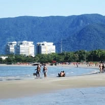 A sunny day at a busy beach with a city skyline in the background. Tall white apartment buildings sit behind a row of palm trees at the water's edge. In the foreground, several people are walking in the shallow water and relaxing on the sand, with a large, blue mountain range rising behind the city.