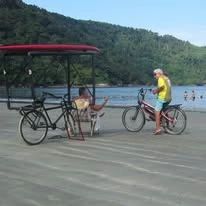 A relaxed beach scene focusing on leisure and transportation. In the foreground, there is a black pedicab bicycle with a covered passenger seat, and next to it stands a person wearing a yellow shirt and blue shorts holding a mountain bike. They are on a sandy beach beside calm water with a forested hill in the distance.