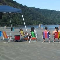 A group of people relaxing on a beach near a calm body of water. They are sitting in colorful folding chairs—blue, red, green, and yellow—under a blue shade canopy, looking out towards the water. A forested hill is visible in the background.