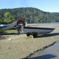 Two small, aluminum motorboats resting on wheeled trailers on a sandy beach. A red all-terrain vehicle (ATV) is hitched to the front of one of the trailers, positioned in front of a bright blue tropical sky and a background of dense, green hills and palm trees.