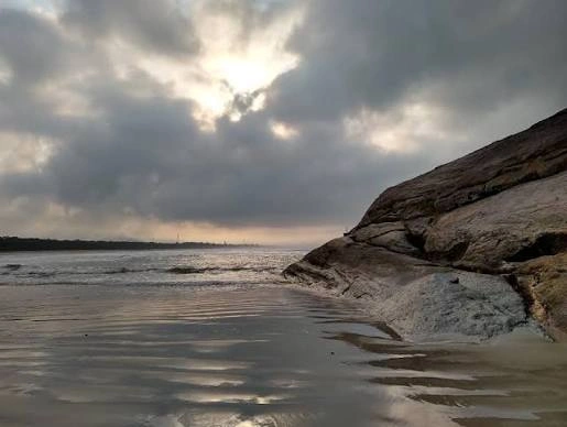 A moody and atmospheric beach scene with large, rough rocks on the right, wet sand reflecting the sky, and a heavy, clouded sky with bright sunbeams breaking through at sunset or sunrise.