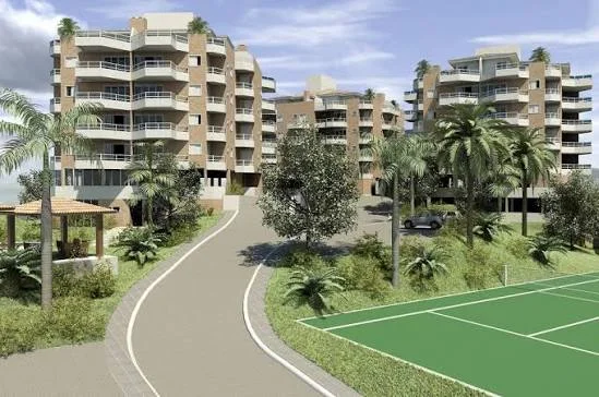 A sunny exterior view of a multi-story residential apartment or resort complex with large balconies, surrounded by palm trees and lush landscaping, featuring a paved driveway and a green tennis court in the foreground.